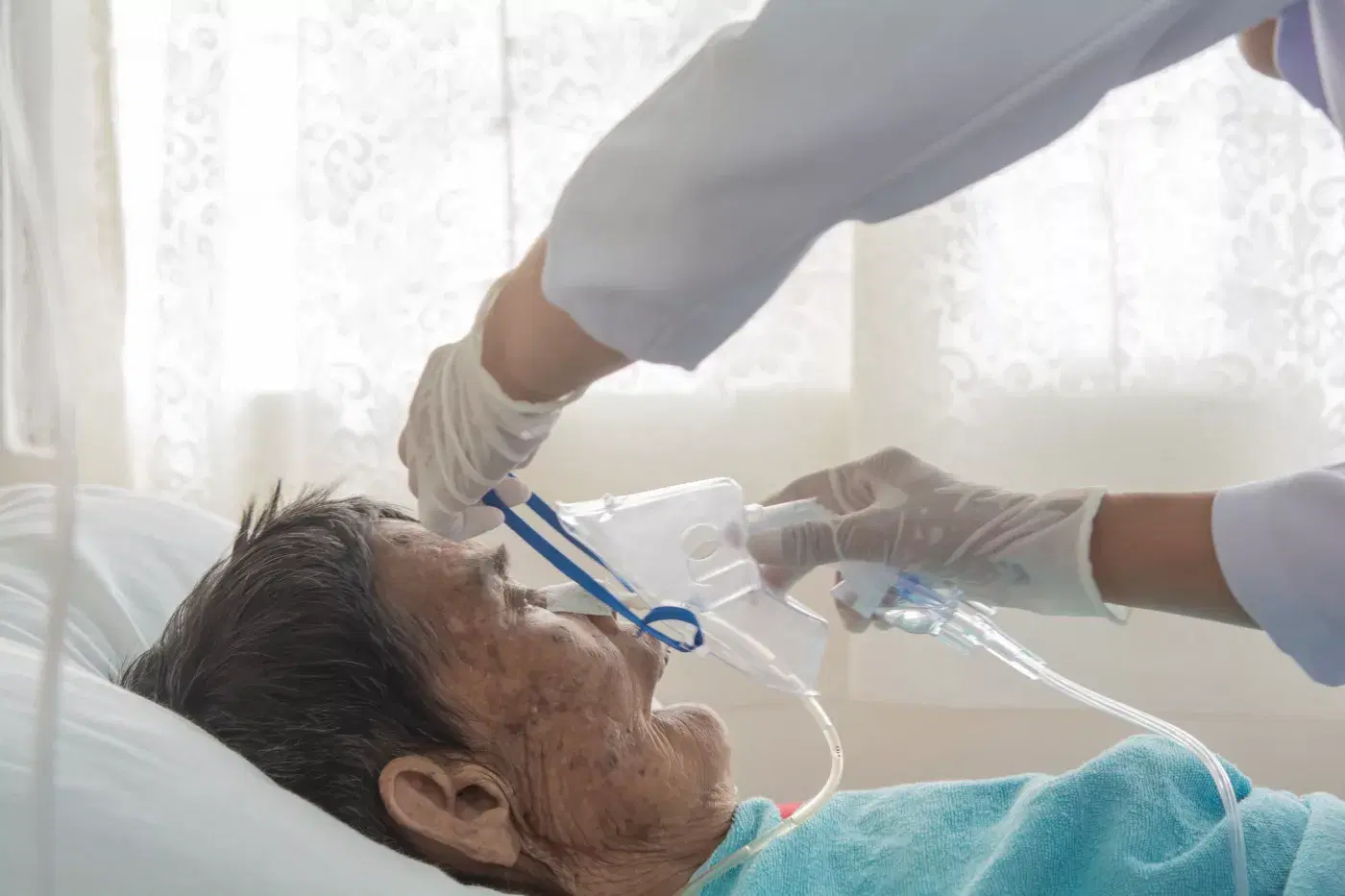 A gloved caregiver fitting an oxygen mask on an elderly bedridden patient at home.