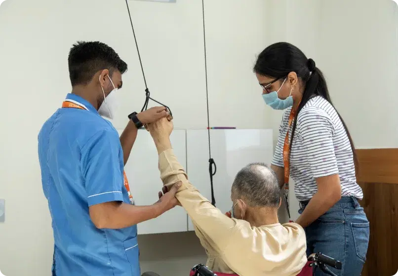 A physiotherapist and nurse assisting a seated patient with overhead arm-lift pulley equipment.